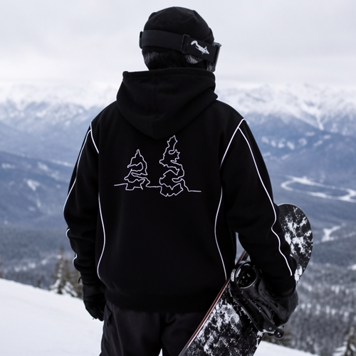 A person standing on a snowy mountain ridge with a snowboard under one arm, wearing a black hooded jacket with a white graphic on the back and dark pants, overlooking a snowy mountain valley.