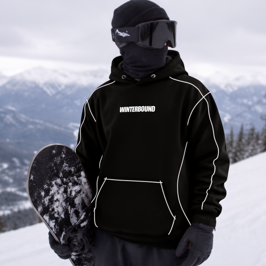 A person standing on a snowy mountain slope wearing a black Winterbound hoodie with white piping, black snowboard pants, a black balaclava, and goggles, holding a snowboard. Ski lifts and mountains are visible in the background.