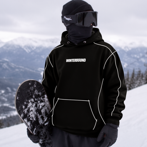 A person standing on a snowy mountain slope wearing a black Winterbound hoodie with white piping, black snowboard pants, a black balaclava, and goggles, holding a snowboard. Ski lifts and mountains are visible in the background.